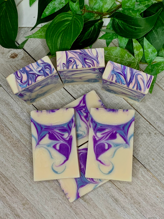 Pictured are 7 bars of Spring in Avalon soap. Four bars of soap are laying face down, three are standing up showing the top design. The top of the soaps show a swirled design of deep purple and shimmery blue on a creamy white background. The same design is featured throughout the bar, as seen on the four bars laying face up.