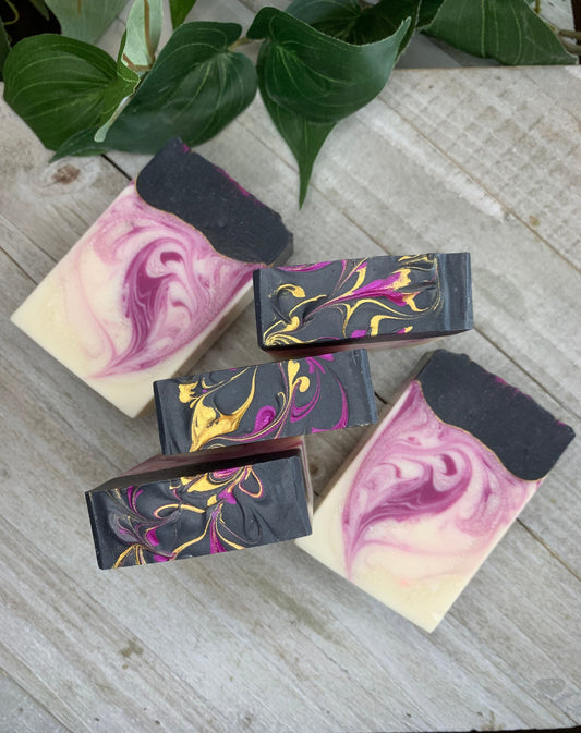 Five bars of Black Raspberry Vanilla scented soap on a wood plank background. Three bars are standing up, showing the top design. The other two soap bars are laid flat to show the inside design. The inside design is three quarters of a raspberry colored swirl in a cream base, and the top quarter of the bar is a black base with raspberry and gold swirls on top.
